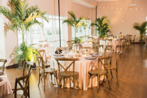 Indoor Wedding Reception with Round Tables and Tropical Palm Trees and String Lights, with Pink Satin Linens, Wooden Crossback Chairs, Vintage Hurricane Lantern Centerpieces | Venue the St Pete Beach Community Center | Tampa Bay Planner UNIQUE Weddings and Events | Rentals A Chair Affair and Over The Top Rental Linens