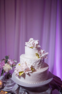 Three Tirered Round White Wedding Cake with Pink Paper Flowers with Gold LEaf Greenery on Gray Cake Stand, with Satin Table LInens White Draping and Purple Uplighting