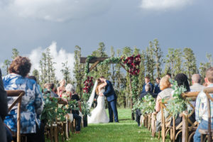 Outdoor Farm Wedding Ceremony First Kiss Portrait, Bride in Sweetheart Strapless Lace Ines Di Santo Dress, Groom in Navy Blue Suit Men's Wearhouse Suit, with Wooden Ceremony Arch with Marsala Red Florals and Greenery, and Wooden Crossback Chairs | Tampa Bay Wedding Planner NK Productions