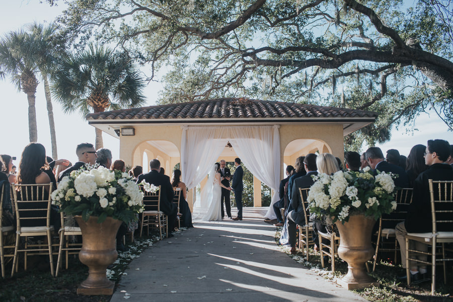Outdoor Southern Waterfront Wedding Ceremony Portrait with Gold Chiavari Chairs, Large Classical Planters with White FLoral and Greenery, and White Draping Ceremony Arch in Gazebo | Bradenton Wedding Venue Powel Crosley Estate | Sarasota Photographer by Brandi Image Photography