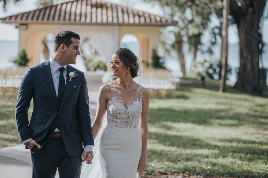Outdoor Courtyard Waterfront Wedding Portrait, Bride with Long Comb Veil in Lace Applique Sweetheart Essence of Australia Dress, Groom in Navy Blue Suit with White Rose Boutonniere | Sarasota Wedding Photographer Brandi Image Photography | Bradenton Venue Powel Crosley Estate