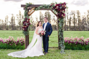 Outdoor Farm Wedding Ceremony Portrait, Bride in Sweetheart Strapless Lace Ines Di Santo Dress with Red Protea and Purple Bouquet, Groom in Navy Blue Suit Men's Wearhouse Suit, with Wooden Ceremony Arch with Marsala and Blush Pink Florals and Greenery | Tampa Bay Wedding Planner NK Productions