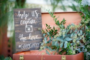 Rustic Outdoor Wedding Reception Instagram Hashtag Sign with White Script handpainted on Natural Wood, with Wild Greenery in Vintage Suitcase