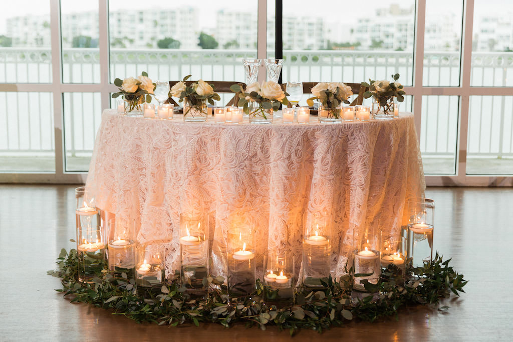 Wedding Reception Sweetheart Table with Textured Blush PInk LInen, Floating Votive Candles in Glass Cylinder Vases, and Small White Roses with Greenery Table Decor and Garland | Waterfront Venue St Pete Beach Community Center | Planner UNIQUE Weddings and Events | Tampa Bay Rentals Over The Top Linen Rentals and A Chair Affair