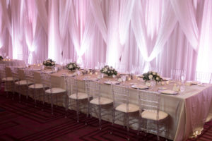 Hotel Ballroom Winter Glam Wedding Reception Long Feasting Table with Champagne Satin Linen, Silver Charger, Clear Chiavari Chairs, and Small White Floral with Greenery Centerpieces and White Draping with Pink Uplighting | Venue Hilton Tampa Downtown | Rentals Kate Ryan Linen Rentals