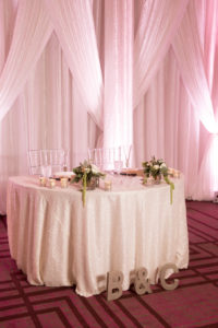 Hotel Ballroom Winter Glam Wedding Reception Sweetheart Table with White Textured Linen, Silver Charger, Clear Chiavari Chairs, and Small White Floral with Greenery Centerpieces and White Draping with Pink Uplighting | Venue Hilton Tampa Downtown | Rentals Kate Ryan Linen Rentals
