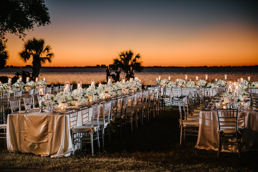 Outdoor Waterfront Sunset Wedding Reception with Long Feasting Tables with Champagne Satin Linens, Low White Floral and Greenery Centerpieces and Silver Candlestickholders and PIllar Candles | Tampa Bay Waterfront Historic Venue Crosley Powel Estate | Satin Linens Over the Top Rental Linens