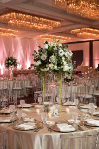 Winter Glam Hotel Ballroom Wedding Reception with Champagne Linens, Clear Chiavari Chairs, and Extra Tall Centerpieces in Geometric Glass Vases with white Floral and Greenery, and White Draping with Blush PInk Uplighting, Silver Chargers | Venue Hilton Tampa Downtown | Rentals Kate Ryan Linen Rentals