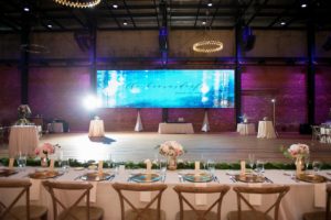 Industrial Chic Wedding Reception with Long Feasting Table with Wooden Cross Back Chairs, Greenery Garland and Small White and Pink Rose Centerpieces in Gold Vases, Gold Chargers, Purple Uplighting and Blue Lighting Projection and Blush and Ivory Linens | Downtown Tampa Wedding Venue Armature Works