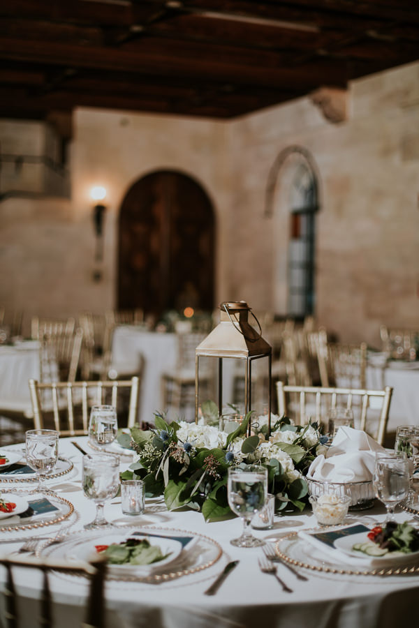 Indoor Wedding Reception with Low White and Greenery Centerpiece with Vintage Hurricane Lantern, Gold Beaded Clear Glass Chargers, Navy BLue Printed Menus, Gold Chiavari CHairs and White Linens | Bradenton Historic Wedding Venue Powel Crosley Estate