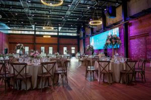 Industrial Chic Wedding Reception with Round Tables with Wooden Cross Back Chairs, Extra Tall and low White and Pink Rose Centerpieces in Gold Vases, Purple Uplighting and Blue Lighting Projection and Blush and Ivory Linens | Exposed Brick Downtown Tampa Wedding Venue Armature Works