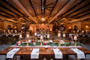 Indoor Rustic Wedding Reception with Wooden Feasting Tables with Greenery Garland Centerpiece and Blush Pink Table Runner, with Marsala Red and BLush Pink Florals, Pillar Candles with Gold Candlestick Holders, White Linens with Rosemary, and String Lights | Tampa Bay Wedding Planner NK Productions