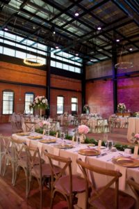Industrial Chic Wedding Reception with Long Feasting Table with Wooden Cross Back Chairs, Greenery Garland and Small White and Pink Rose Centerpieces in Gold Vases, Gold Chargers, Purple Uplighting and Blush and Ivory Linens | Downtown Tampa Wedding Venue Armature Works