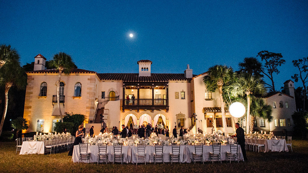 Outdoor Waterfront Sunset Wedding Reception with Long Feasting Tables with Champagne Satin Linens, Low White Floral and Greenery Centerpieces and Silver Candlestickholders and PIllar Candles | Tampa Bay Waterfront Historic Venue Crosley Powel Estate | Satin Linens Over the Top Rental Linens