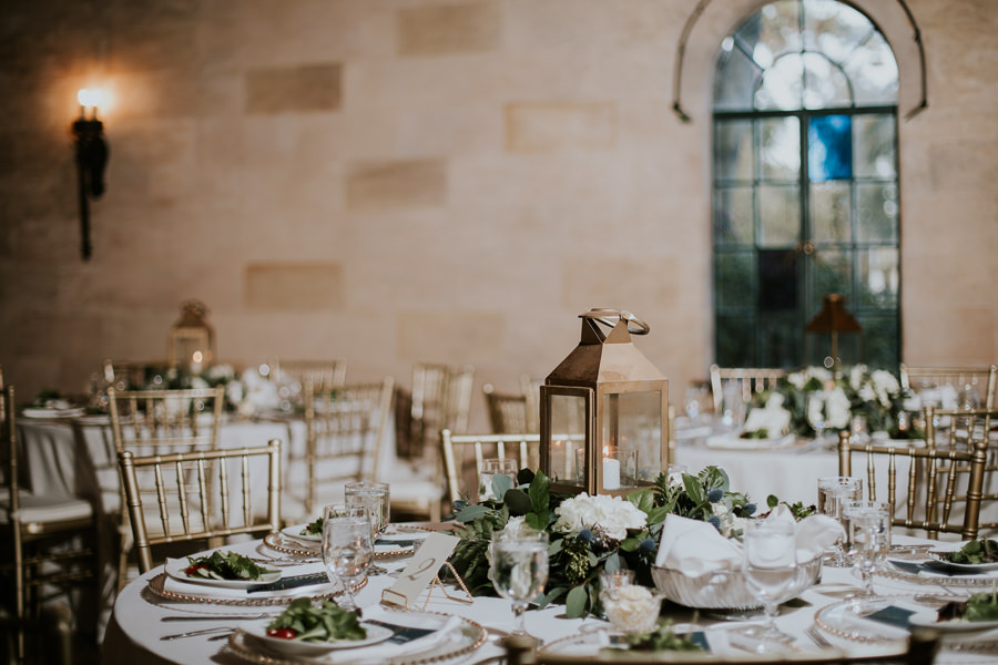 Indoor Wedding Reception with Low White and Greenery Centerpiece with Vintage Hurricane Lantern, Gold Beaded Clear Glass Chargers, Navy BLue, White and Gold Printed Menus, Gold Chiavari CHairs and White Linens | Bradenton Historic Wedding Venue Powel Crosley Estate