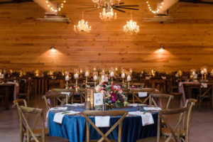 Indoor Rustic Wedding Reception with Round Table with Navy Blue Tablecloth, withMedium Height Centerpiece with Marsala Red and BLush Pink Protea and Florals, Pillar Candles with Gold Candlestick Holders, White Linens with Rosemary, and Navy Blue Printed on White Table Number in Tree Bark Stand, Wooden Cross Back Chairs and Hanging Chandeliers and String Lights | Tampa Bay Wedding Planner NK Productions