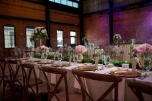 Industrial Chic Wedding Reception with Long Feasting Table with Wooden Cross Back Chairs, Greenery Garland and Small White and Pink Rose Centerpieces in Gold Vases, Gold Chargers, Purple Uplighting and Blush and Ivory Linens | Downtown Tampa Wedding Venue Armature Works