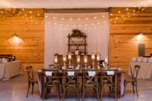 Indoor Rustic Wedding Reception with Wooden Feasting Tables with Greenery Garland Centerpiece and Blush Pink Table Runner, with Marsala Red and BLush Pink Florals, Pillar Candles with Gold Candlestick Holders, White Linens with Rosemary, and String Lights with Cross Back Chairs | Tampa Bay Wedding Planner NK Productions