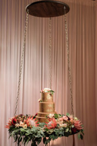 Four Tiered Round Copper and Gold Wedding Cake with Marsala Red Protea, Blush PInk and Cream Florals with Greenery, on Round Wooden Suspended Platform Table Hanging from Chains | Tampa Bay Wedding Planner NK Productions