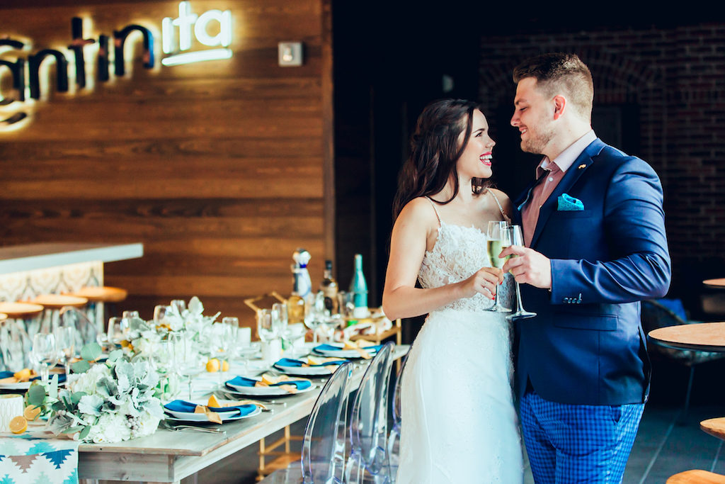 Industrial Chic Intimate Restaurant Bar Wedding Reception Portrait, Bride in Belted Column Dress with Lace Spaghetti Strap Bodice, Groom in Blue Suit with Pink Shirt and Checked Pants, Long Wooden Feasting Table with Turquoise and Linens with Cut Lemon Accents, Low Greenery Centerpiece, and Gray and Green Woven Canvas Runner | Modern Downtown St Pete Cantina Wedding Venue Red Mesa Events | Tampa Bay Wedding Planner Jennifer Matteo Event Planning