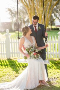 Outdoor First Look Portrait on Swing, Bride in Illusion Lace V Neck Paloma Blanca Dress with Sage Greenery, Pink, Burgundy, Black and White Floral Bouquet, Groom in Gray Suit with Succulent Greenery Boutonniere and Red TIe | Sarasota Wedding Photographer Andi Diamond Photography