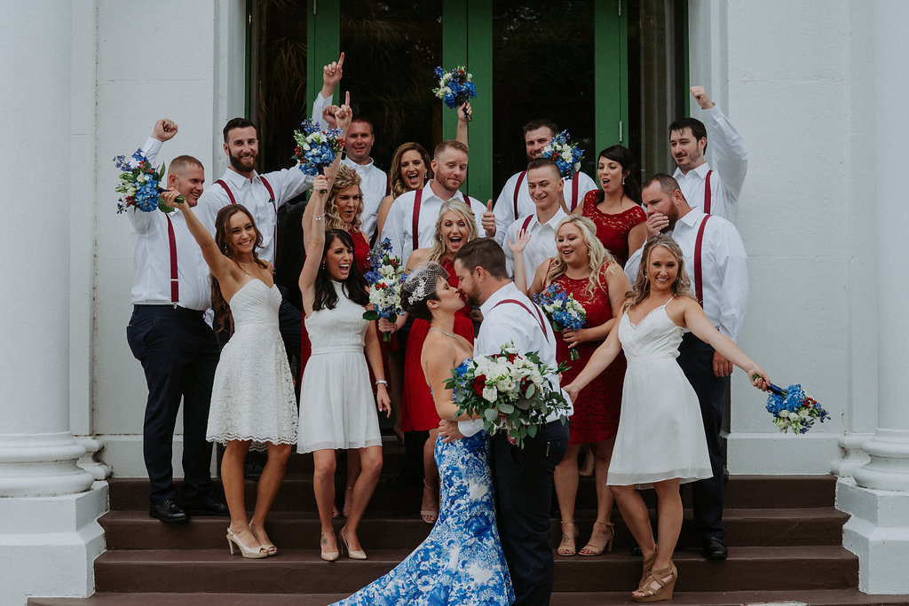 Outdoor Church Steps Wedding Party Portrait, Groom and Groomsmen in White Shirts with Navy Pants and Red Suspenders, Bridesmaids in Mismatched Short Red and White Dresses, Bride in Strapless Mermaid BLue and White Printed Dress, with White, Blue and Red Floral with Greenery Bouquet | Tampa Bay Wedding Photographer Grind and Press Photography | St Pete Hair and Makeup Michele Renee The Studio | Florist Apple Blossoms Floral Designs