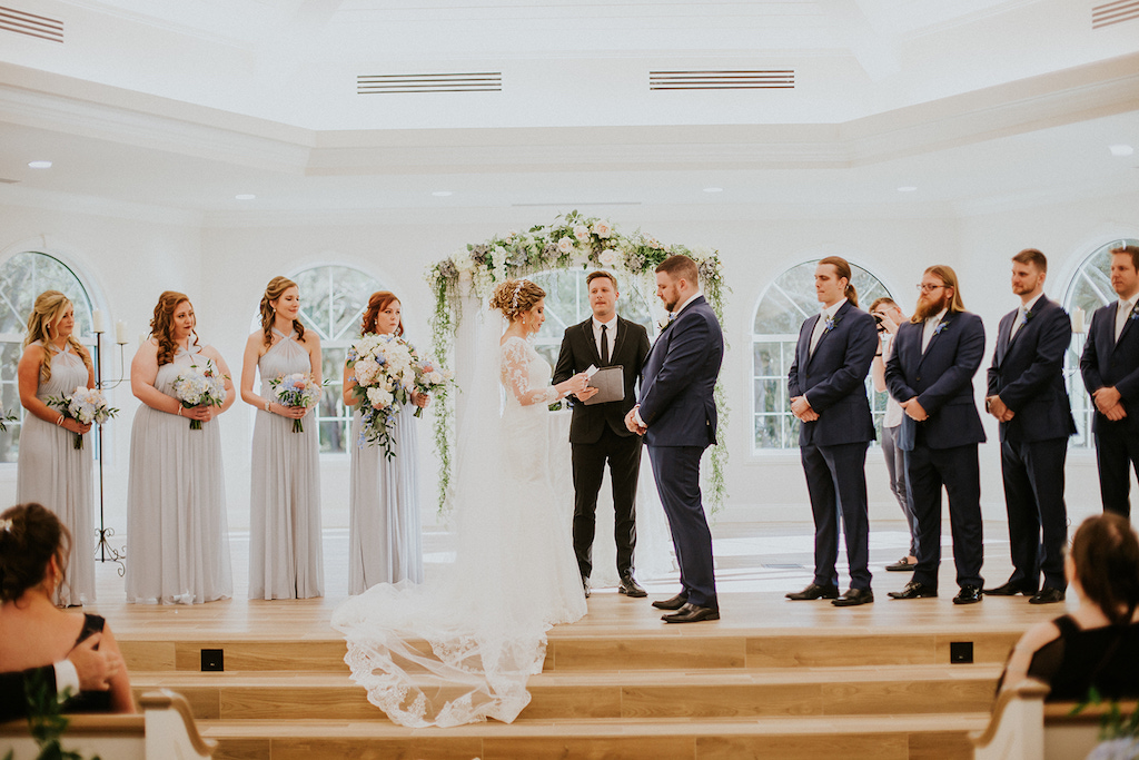 Indoor Wedding Ceremony Portrait, Groomsmen Wearing Navy Blue Suits with Blue Flower Boutonniere, Bridesmaids Wearing Silver/Grey Dresses with White and Greenery Bouquets, Bride Wearing White Lace Wedding Dress with Cathedral Length Lace and Tulle Veil and Groom Wearing Navy Blue Suit | Safety Harbor Wedding Venue Harborside Chapel