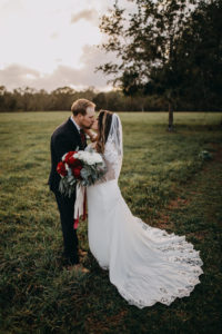 Outdoor Sunset Bride and Groom Wedding Portrait, Groom Wearing Navy Blue Suit with Grey Vest, Red Tie and White Floral Boutonniere, Bride in Lace David's Bridal Wedding Dress, and Red Peonies, White Roses and Greenery Bouquet with Red and White Ribbon, and Lace Veil | Tampa Bay Wedding Photographer Rad Red Creative | Lithia Rustic Wedding Venue Southern Grace