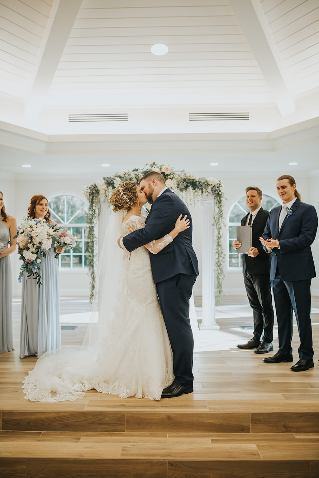 Wedding Ceremony Portrait, Groomsmen Wearing Navy Blue Suits with Blue Flower Boutonniere, Bridesmaids Wearing Silver/Grey Dresses with White and Greenery Bouquets, Bride Wearing White Lace Wedding Dress with Cathedral Length Lace and Tulle Veil and Groom Wearing Navy Blue Suit | Safety Harbor Wedding Venue Harborside Chapel