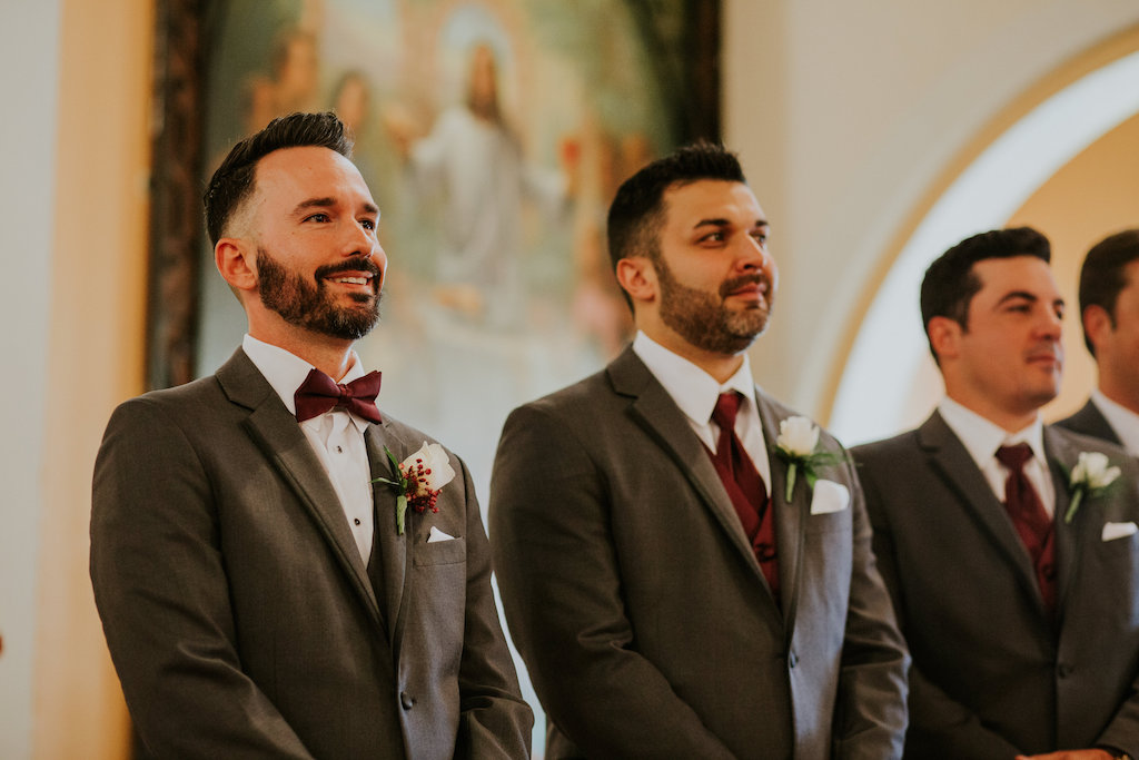 Groom's Reaction to Bride Waking Down the Aisle Wedding Ceremony Portrait in Grey Tuxedos and Burgundy Bowties with White Rose and Red Flowers with Greenery Boutonniere | Tampa Bay Wedding Venue Our Lady of Perpetual Help Catholic Church