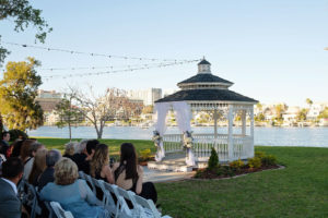 Outdoor Garden Wedding Ceremony Decor with White Wooden Folding Chairs, White and Greenery Floral Decor and Waterside Gazebo with White Draping and Bistro Hanging Lights | Tampa Wedding Ceremony Venue Davis Island Garden Club | Tampa Bay Wedding Photographer Marc Edwards Photographs | String Market Lights Gabro Event Services