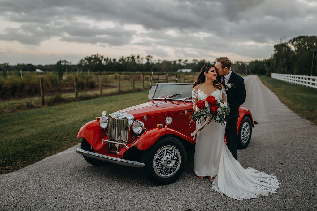 Outdoor Bride and Groom Wedding Portrait, Groom Wearing Navy Blue Suit with Grey Vest, Red Satin Tie and White Flower and Greenery Boutonniere, Bride in Long Sleeve Lace David's Bridal Wedding Dress, and Red Peonies, White Roses and Greenery Bouquet in Vintage Red Convertible Car | Tampa Bay Wedding Photographer Rad Red Creative | Lithia Rustic Wedding Venue Southern Grace