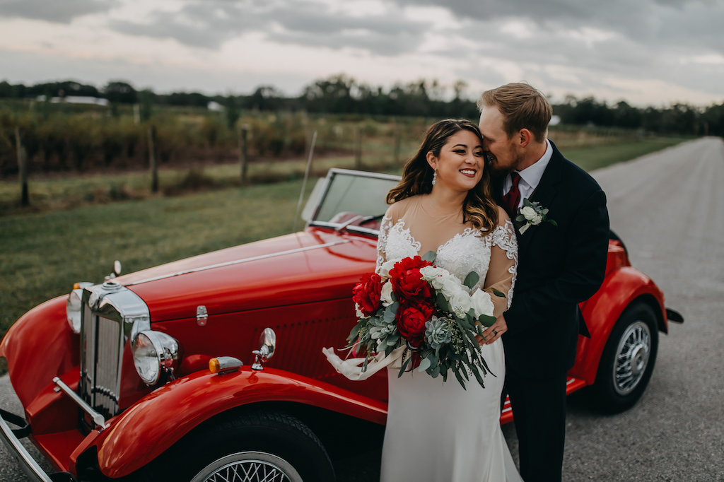 Outdoor Bride and Groom Wedding Portrait, Groom Wearing Navy Blue Suit with Grey Vest, Red Satin Tie and White Flower and Greenery Boutonniere, Bride in Long Sleeve Lace David's Bridal Wedding Dress, and Red Peonies, White Roses and Greenery Bouquet in Vintage Red Convertible Car | Tampa Bay Wedding Photographer Rad Red Creative | Lithia Rustic Wedding Venue Southern Grace