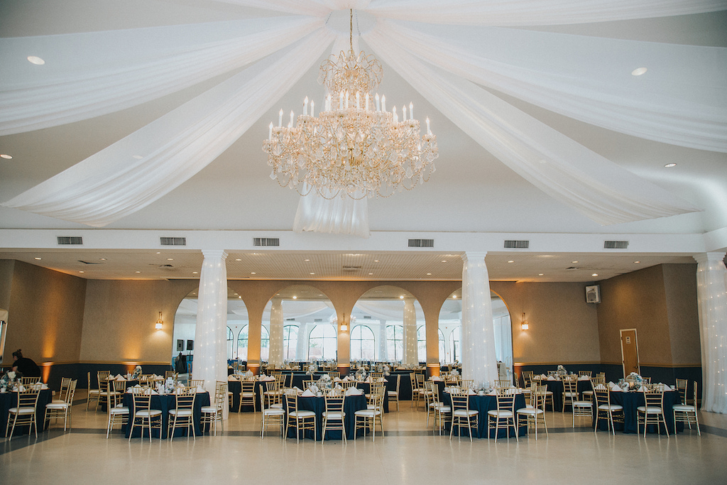 Navy Blue, Gold and Blush Wedding Reception Hall with Gold Chandelier, Navy Blue Table Cloths/Linens, and Gold Chiavari Chairs and White Drapery | Clearwater Wedding Reception Venue Matheos Hall