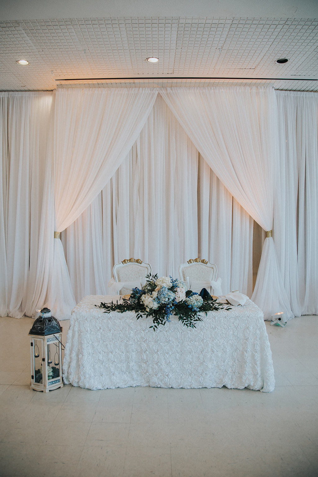 Indoor Wedding Reception Hall Sweetheart Table with White Textured Linen, Fancy Chairs, White Lantern, and White, Blue and Blush Floral with Greenery Centerpieces and White Draping with Off-White Uplighting