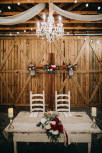 Indoor Barn Reception Decor with Crystal Chandelier, String Lights, Drapery, Bride and Groom Sweetheart Table with Wooden Farm Table with Red Peonies, White Roses and Greenery Bouquet, Silver Pedestals with White Pillar Candles, White Wooden Chairs, Wooden Cross with Red, White and Greenery Bouquets | Tampa Bay Wedding Photographer Rad Red Creative | Lithia Wedding Venue Southern Grace