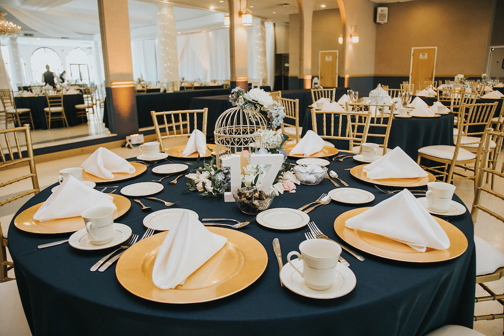 Navy Blue, Gold and Blush Wedding Reception Hall with Navy Blue Table Cloths/Linens, and Gold Chiavari Chairs, Gold Plate Chargers, Birdcage Centerpiece with White, Blue and Blush Florals and Greenery and White Wooden Table Number and White Hanging Drapery | Clearwater Wedding Reception Venue Matheos Hall
