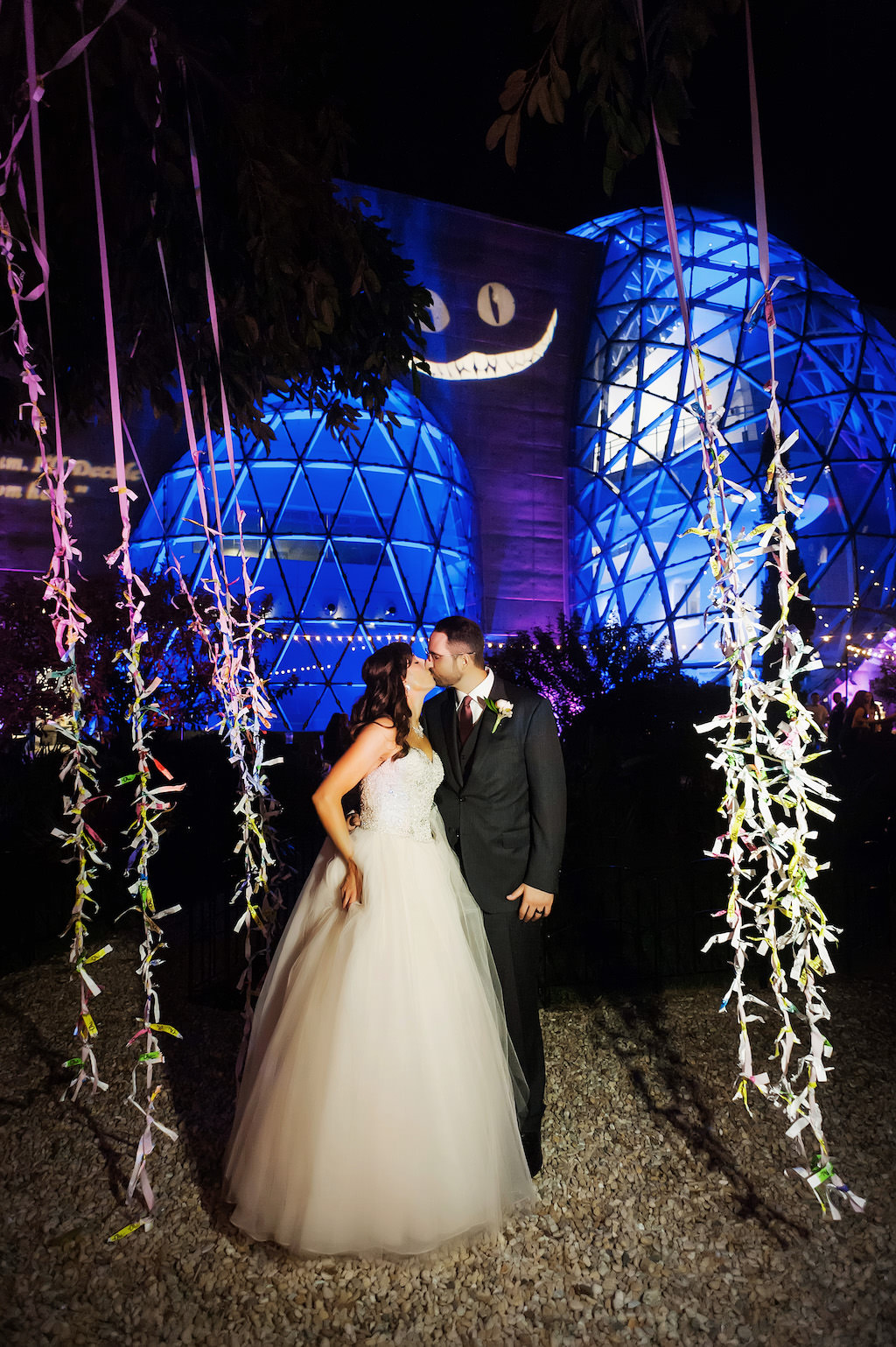 Bride and Groom Kissing Surrounded by Hanging Branches with Leaves in Blush Pink Wedding Dress with Tulle Bottom in Front of Blue Uplighted Dali Museum Decorated with Alice in Wonderland Cheshire Cate Face | Tampa Bay Bridal Shop Truly Forever Bridal