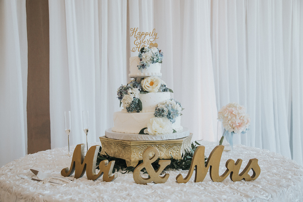 White Wedding Cake with White and Blue Flowers, Gold Cake Topper on Top Antique Cake Stand and Greenery, Champagne Flutes, and Gold Mr and Mrs Wooden Cutout Sign on Textured White Linen