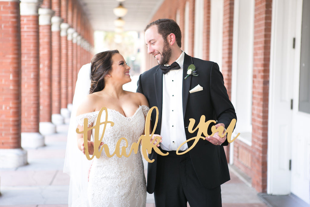 Outdoor Wedding Portrait with Stylish Gold Oversized Letter Thank You Sign, Bride in Off The Shoulder Lace Augusta Jones Dress, Groom in Black Tuxedo with Bow Tie, Blush Pink Pocket Square and Rose Boutonniere with Greenery | Tampa Bay Wedding Photographer Carrie Wildes Photography