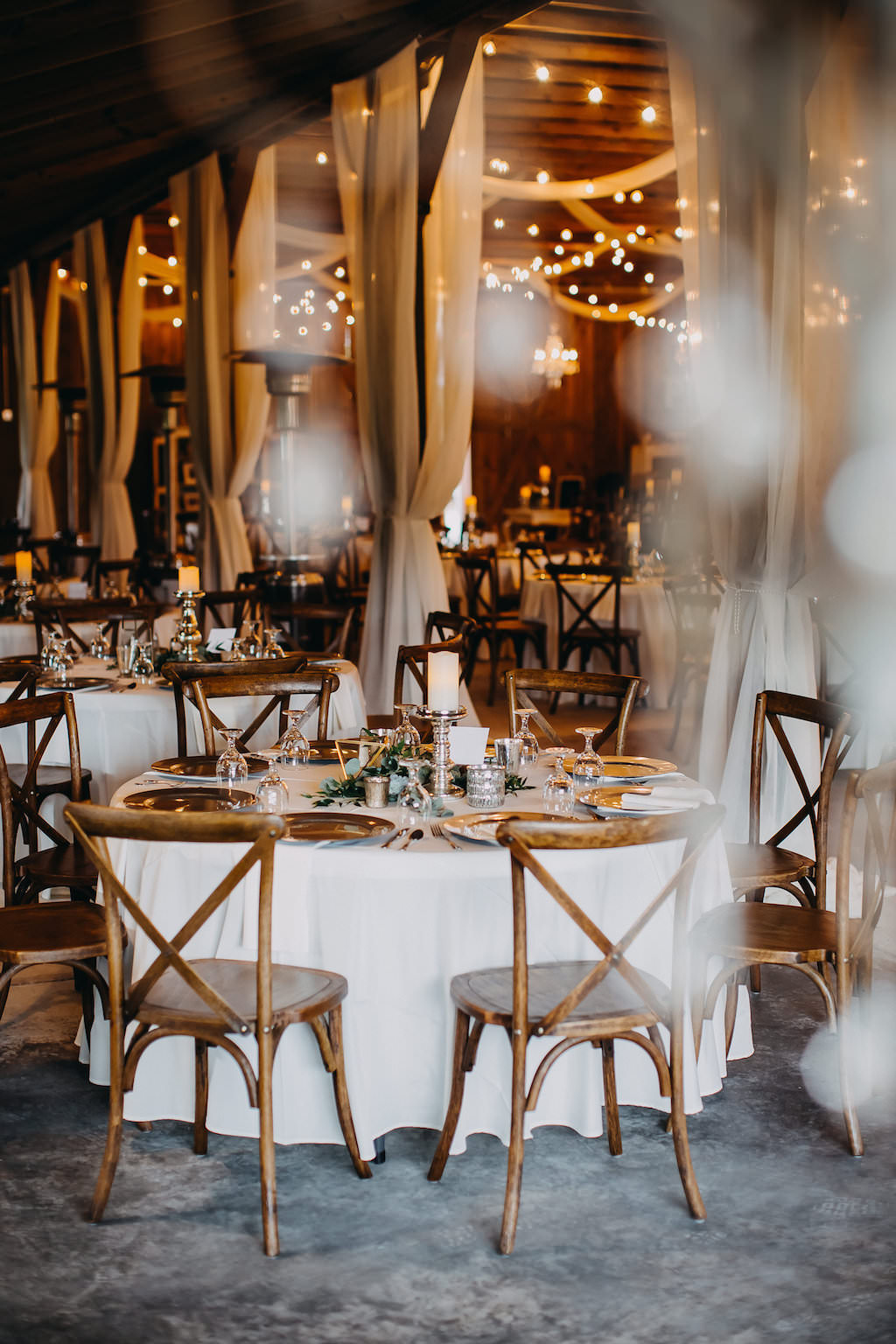 Indoor Rustic Barn Wedding Reception Decor with Round Table with White Tablecloth, with Pillar Candles with Silver Candlestick Holders Centerpieces, Greenery, Wooden Cross Back Chairs and String Lights and Drapery | Tampa Bay Wedding Photographer Rad Red Creative | Lithia Wedding Venue Southern Grace