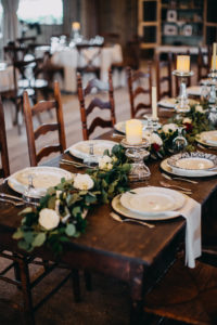 Indoor Rustic Barn Wedding Reception Decor with Long Wooden Feasting Tables with Greenery Garland Centerpiece and White Roses with Pillar Candles with Silver Candlestick Holders, White Linens with Decorative China Plate Setting | Tampa Bay Wedding Photographer Rad Red Creative | Lithia Wedding Venue Southern Grace