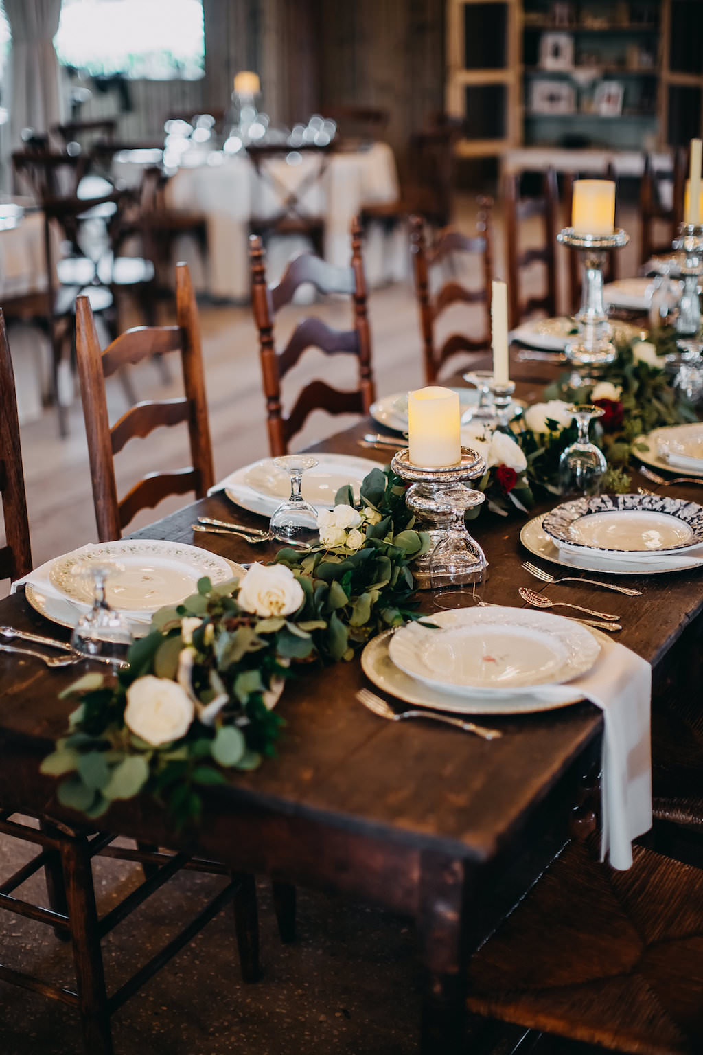 Indoor Rustic Barn Wedding Reception Decor with Long Wooden Feasting Tables with Greenery Garland Centerpiece and White Roses with Pillar Candles with Silver Candlestick Holders, White Linens with Decorative China Plate Setting | Tampa Bay Wedding Photographer Rad Red Creative | Lithia Wedding Venue Southern Grace