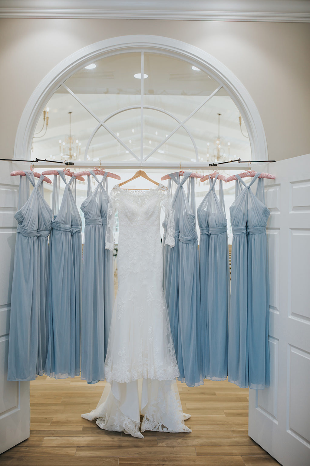 White Stella York Lace Wedding Dress with Long Sleeves Hanging on Wooden Hanger in Venue and Light Blue/Dusty Blue Chiffon Bridesmaid Dresses Hanging in Venue
