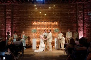 Bride and Groom Wedding Ceremony Portrait, Exposed Brick and Industrial String Lights, White Pedestals with Glass Cylinders and Baby's Breathe | Modern Industrial Wedding Reception Decor Inspiration | Downtown St. Petersburg Wedding Venue NOVA 535 | St. Pete Photographer Marc Edwards Photography