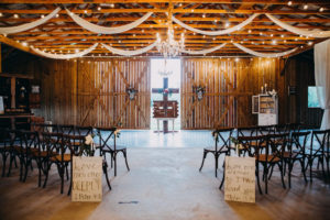 Elegant, Romantic Barn Wedding Ceremony Decor with String Market Lights, Ceiling Draping and Chandelier, Wooden French Country Chairs, Wooden Cross with Red, White and Greenery Bouquet and Wedding Aisle Signage | Tampa Bay Wedding Photographer Rad Red Creative | Lithia Rustic Wedding Venue Southern Grace