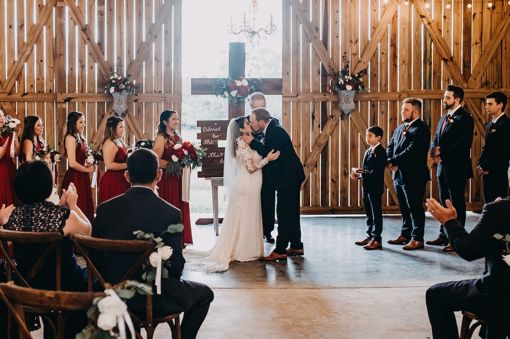 Barn Wedding Reception, Wooden Cross with Red, White and Greenery Bouquet, Bride and Groom Wedding First Kiss Portrait, Bride Wearing White Long Sleeve Lace Wedding Dress and Veil, Groom Wearing Navy Blue Suit, Bridesmaids in Long Burgundy Dresses with Straps Holding Red and White Bouquets, Groomsmen Wearing Navy Blue Suits and Red Ties with Red and White Floral Boutonnieres | Tampa Bay Wedding Photographer Rad Red Creative | Lithia Rustic Wedding Venue Southern Grace