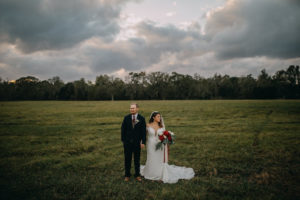 Outdoor Bride and Groom Wedding Portrait, Groom Wearing Navy Blue Suit with Grey Vest, Red Tie and White Floral Boutonniere, Bride in White Long Sleeve Lace David's Bridal Wedding Dress, and Red Peonies, White Roses and Greenery Bouquet with Red and White Ribbon, and Lace Veil | Tampa Bay Wedding Photographer Rad Red Creative | Lithia Rustic Wedding Venue Southern Grace