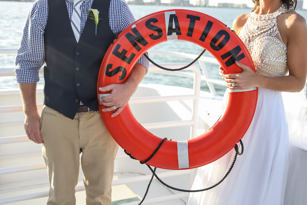 Outdoor Waterfront Bride and Groom Portrait Holding Lifesaver Tube, Bride Wearing Nude Beaded Scoop Neck and White Chiffon Floor Length Wedding Dress, Groom Wearing Khaki Pants with Blue and White Checkered Dress Shirt and Blue and White Striped Tie with Navy Blue Vest and Ivory Calla Lilly Boutonniere | Tampa Bay Nautical Wedding Venue Yacht Starship | Tampa Bay Wedding Photographer Lifelong Photography Studios