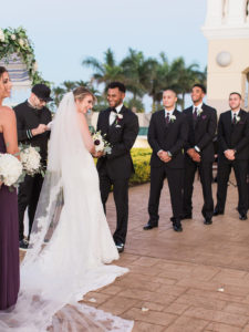 Outdoor Wedding Ceremony Portrait, Bride in Illusion Spaghetti Strap Illusion Lowback Lace Dress and Cathedral Long Veil, Groom in Black Tuxedo and Red and White Flower Boutonniere, Groomsmen in Black Tuxedos and Deep Purple Ties with White Flower Boutonniere, Greenery and White Rose Arch | Clearwater Beach Wedding Venue Feather Sound Country Club